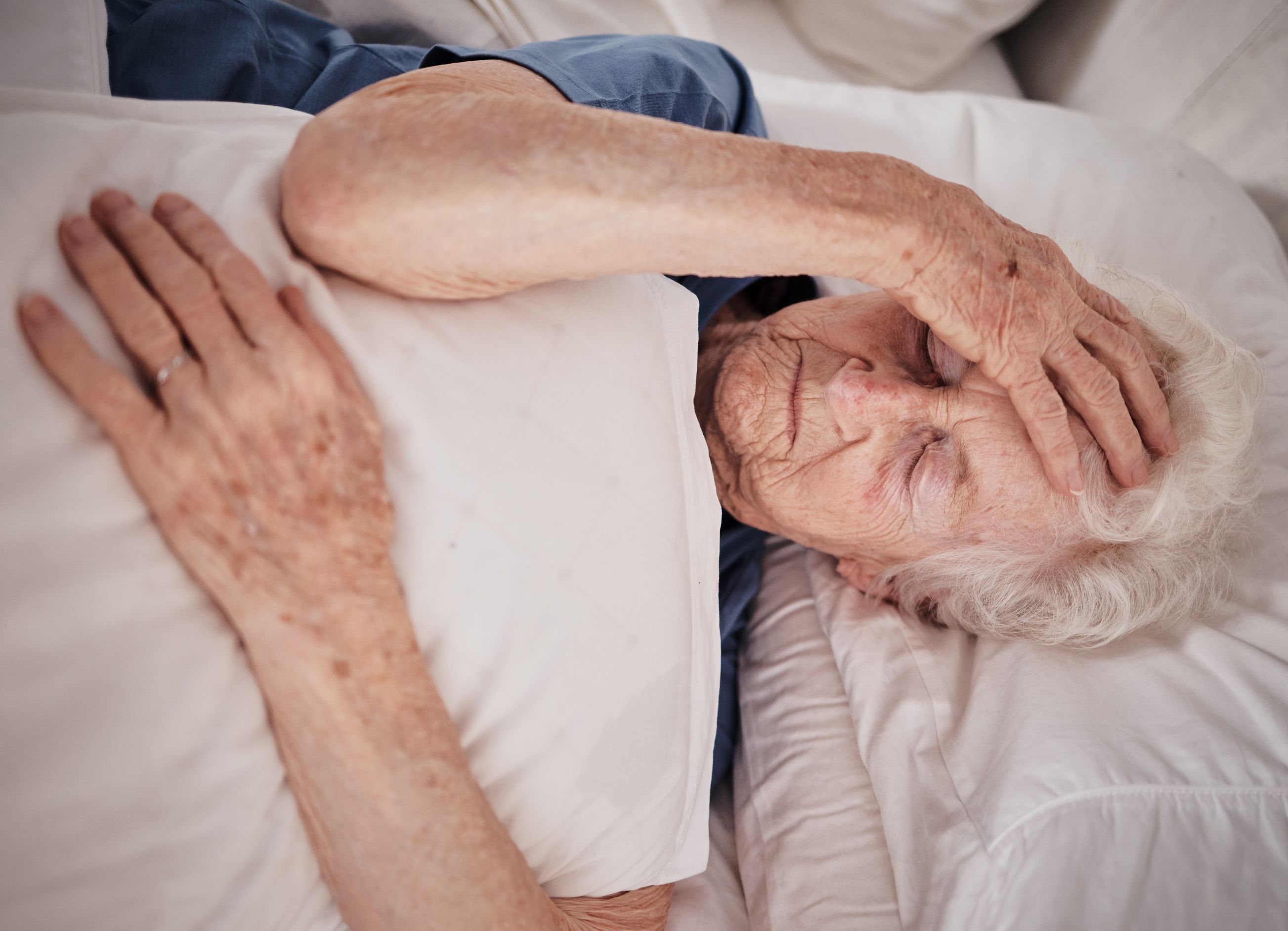 Elder woman lies in nursing home bed holding her forehead with one hand as if in pain.